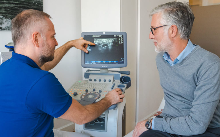 Münchener Orthopäde bei der Arbeit mit einem älteren männlichen Patienten, der ihm etwas auf einem Display zeigt