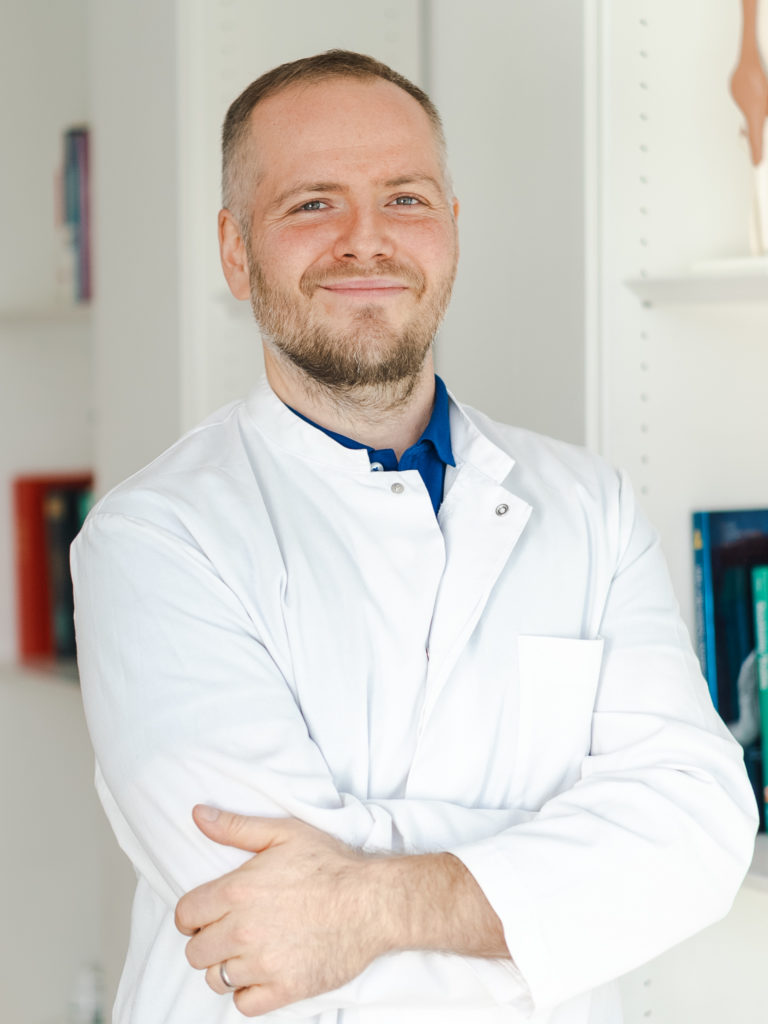 Dr. Michael Wunderlich, ein Orthopäde aus München in einem weißen Arztkittel, schaut lächelnd in die Kamera.