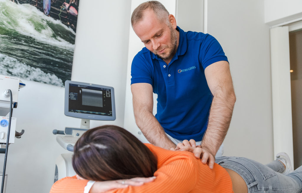 Dr. Michael Wunderlich behandelt als Osteopathe in München eine Klientin im orangefarbenen Hemd. Sie liegt auf einem Tisch auf dem Bauch.