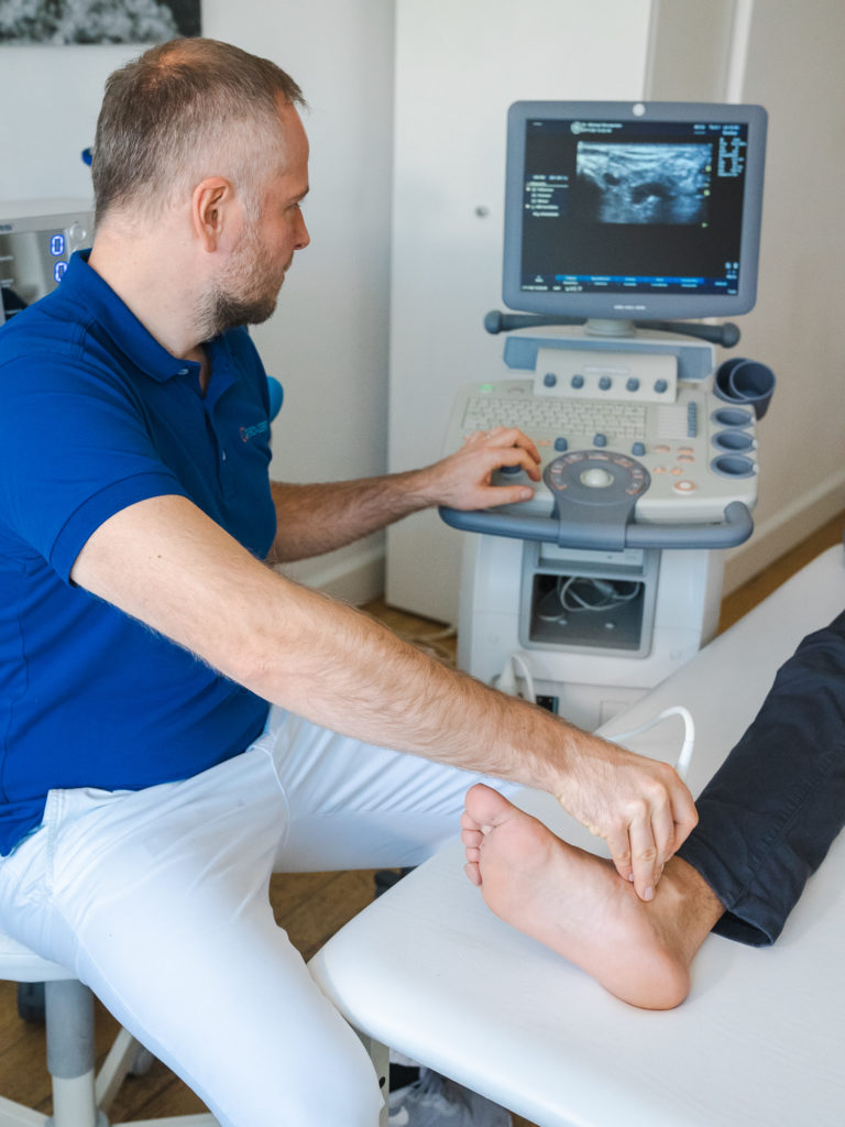 Fußspezialist in München, der mit einem Untersuchungsgerät arbeitet, während der Patient auf einem Bett liegt und einer seiner Füße sichtbar ist.
