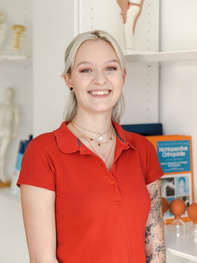 Eine Mitarbeiterin in der orthopädischen Praxis Ortho-Lehel München, die ein rotes Hemd trägt, lächelt in die Kamera und hat eine Tätowierung auf dem linken Arm.