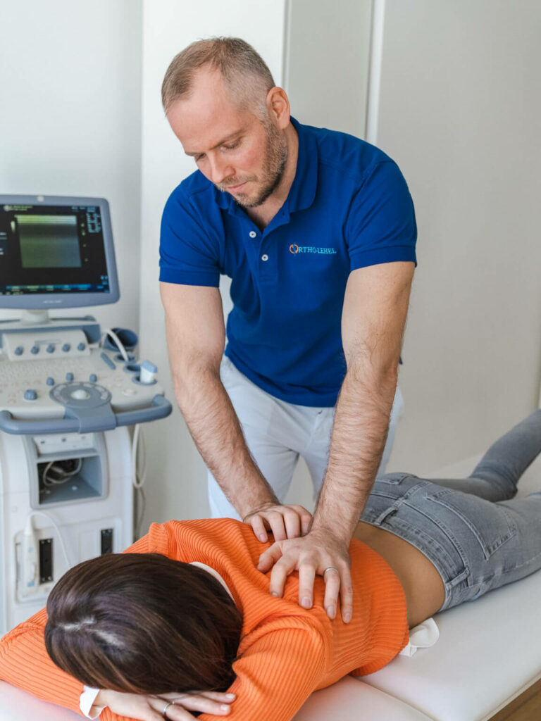 Orthopäde München bei der Behandlung einer Patientin im orangefarbenen Hemd, die auf einem Tisch liegt